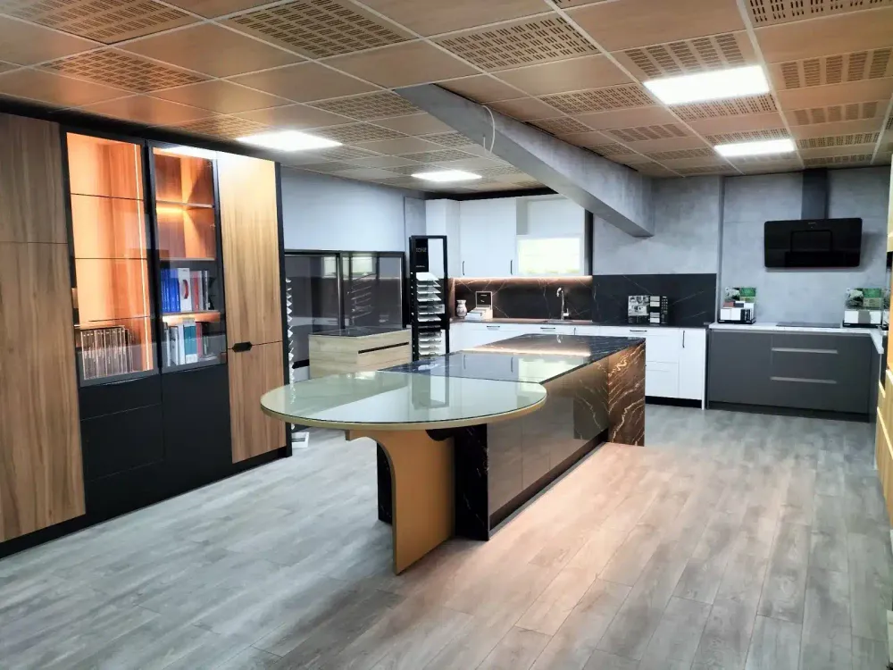 Isla con encimera de piedra oscura veteada, mesa redonda de cristal y columnas de madera con vitrinas iluminadas en la exposición de Kitchenmania